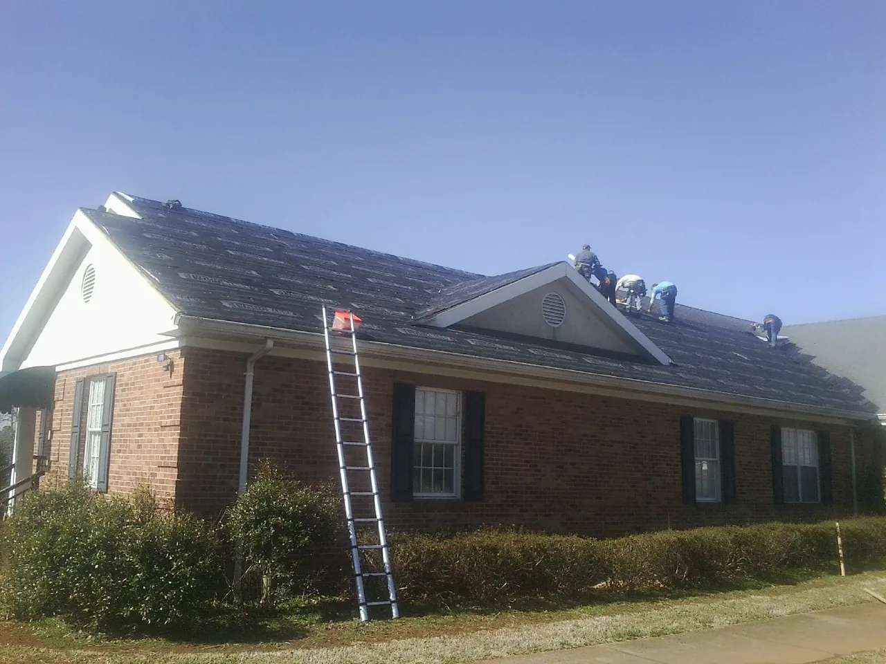 Crew tearing shingles off a brick ranch roof with a ladder set — Metal Roof Repair work by our crew in East Templeton