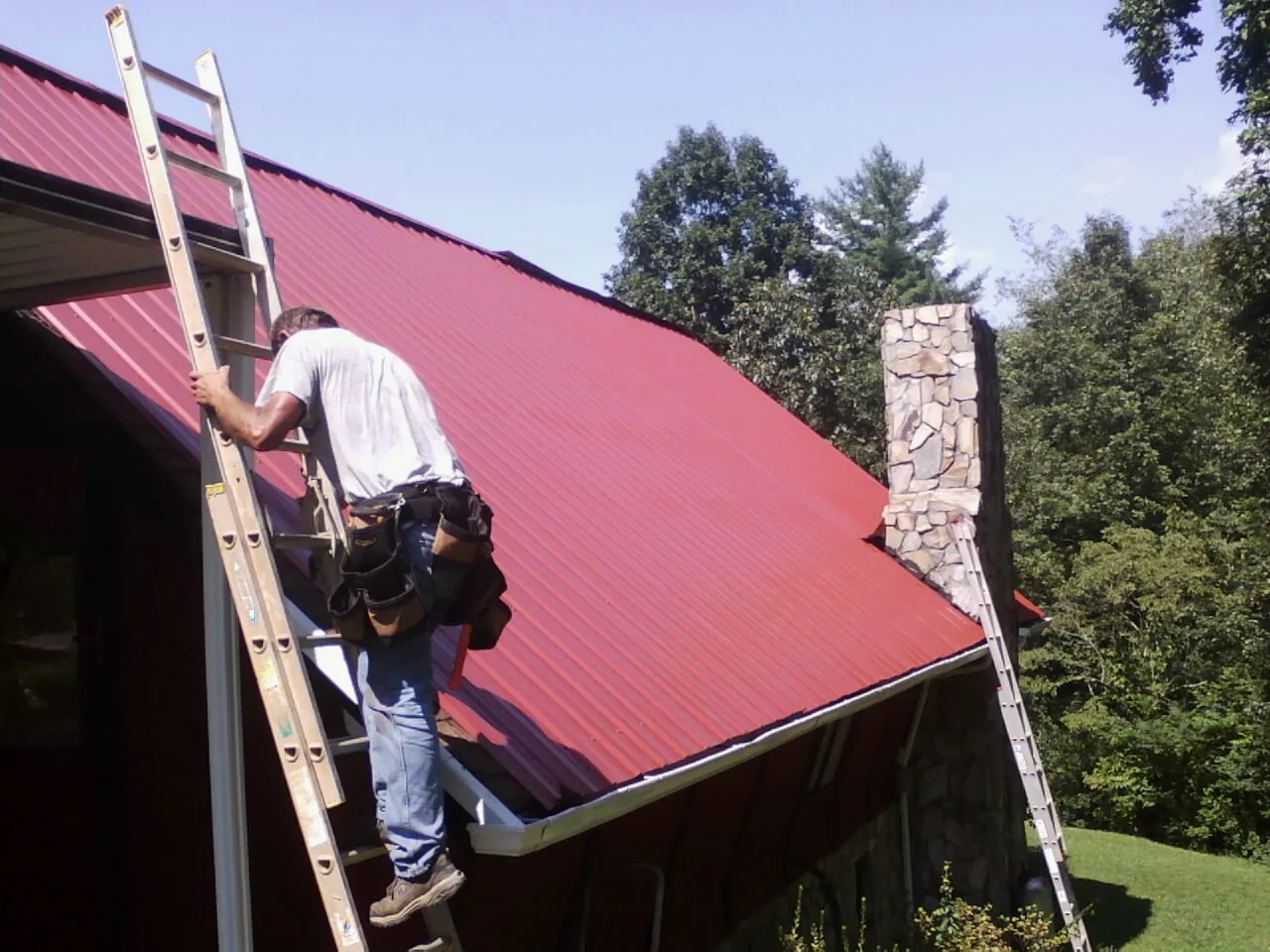 Roofer on a ladder beside a red metal roof and stone chimney — Soffit & Fascia Repair by Roofing Brothers in East Templeton